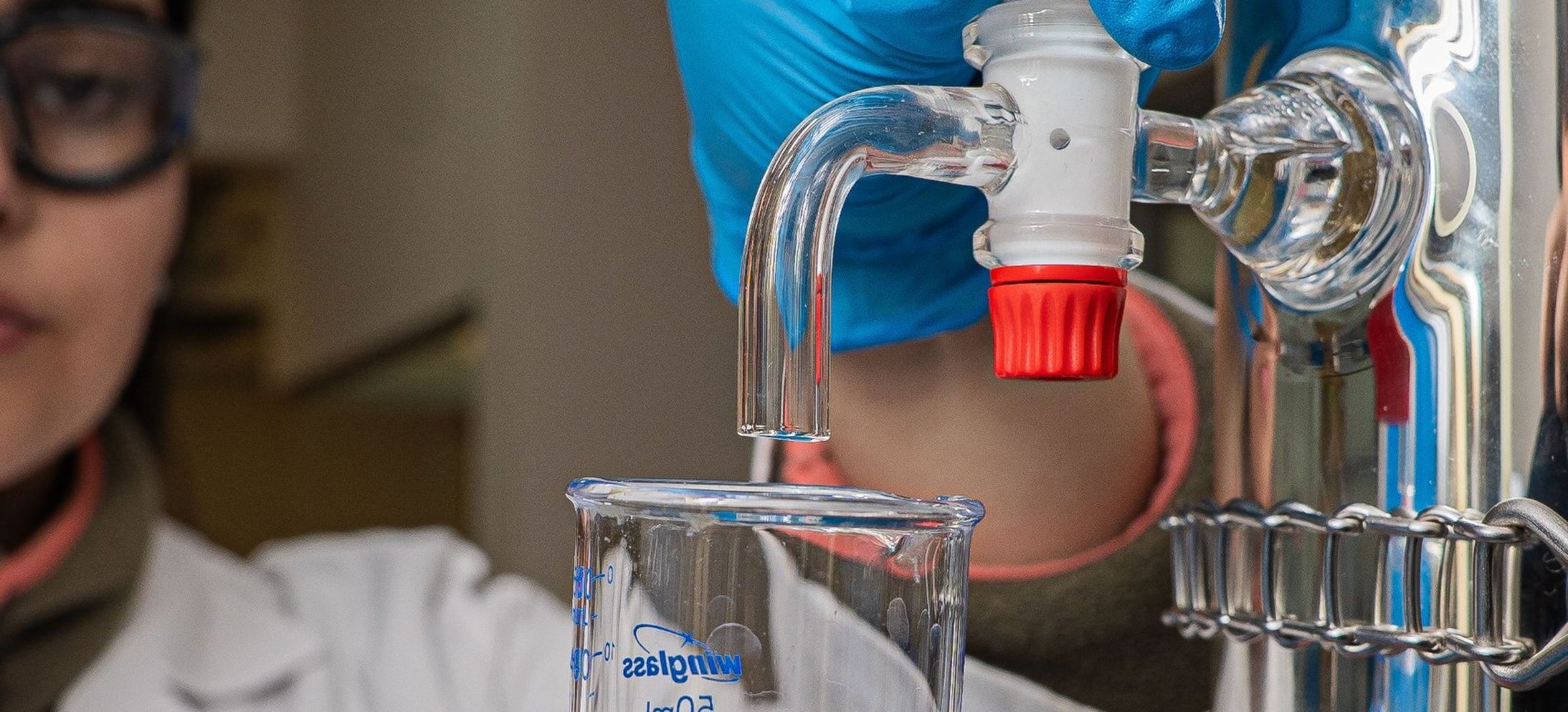 Gloved hands collecting liquid from a glass tap into a beaker, a scientist in safety goggles behind