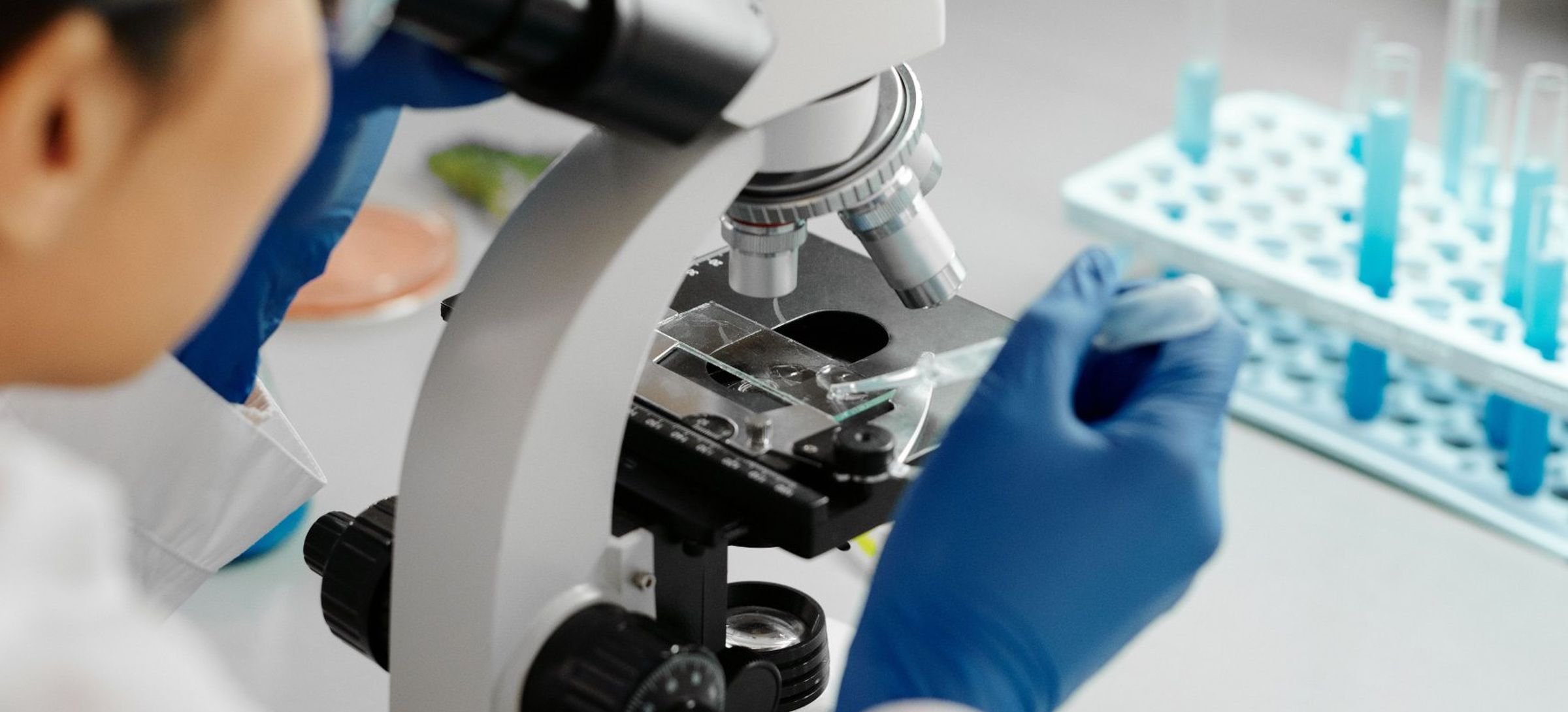A researcher in gloves placing a slide under a microscope, with a rack of blue sample tubes beside them