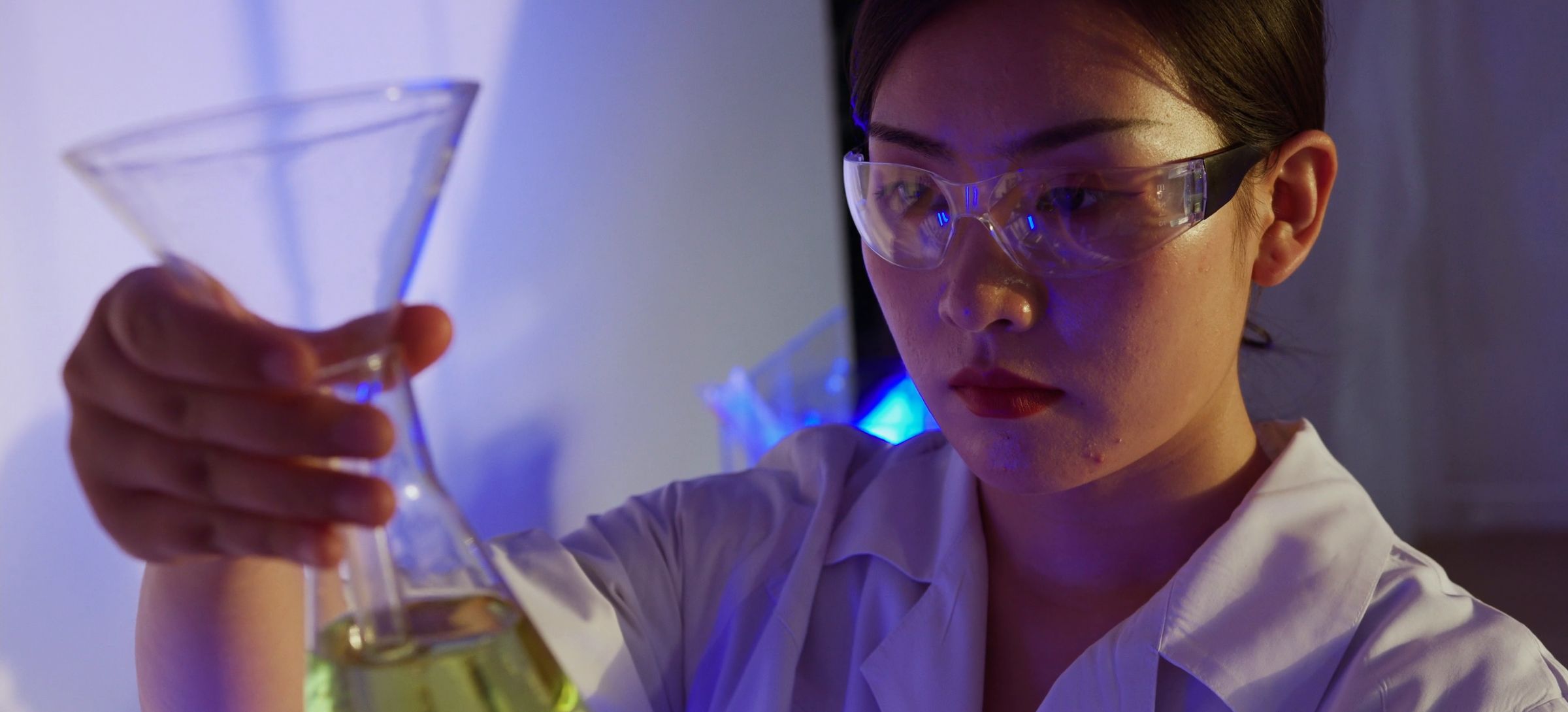 A scientist in safety glasses examining a flask of liquid under purple lab light